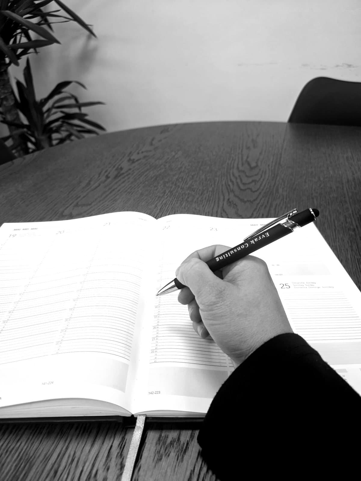 Zwart-witfoto van een hand die met een pen van Evrak Consulting in een open agenda schrijft.