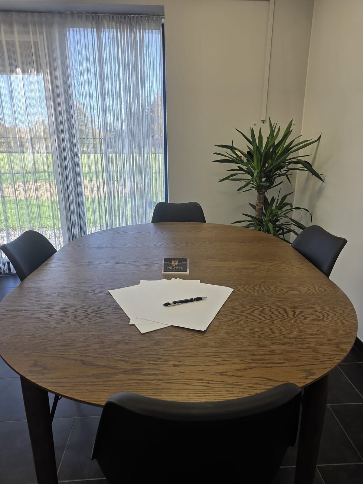 De vergadertafel van het kantoor: een ronde houten tafel met blanco papier, een pen en een kaarthouder van Evrak Consulting, bij een raam met zicht op een grasveld.